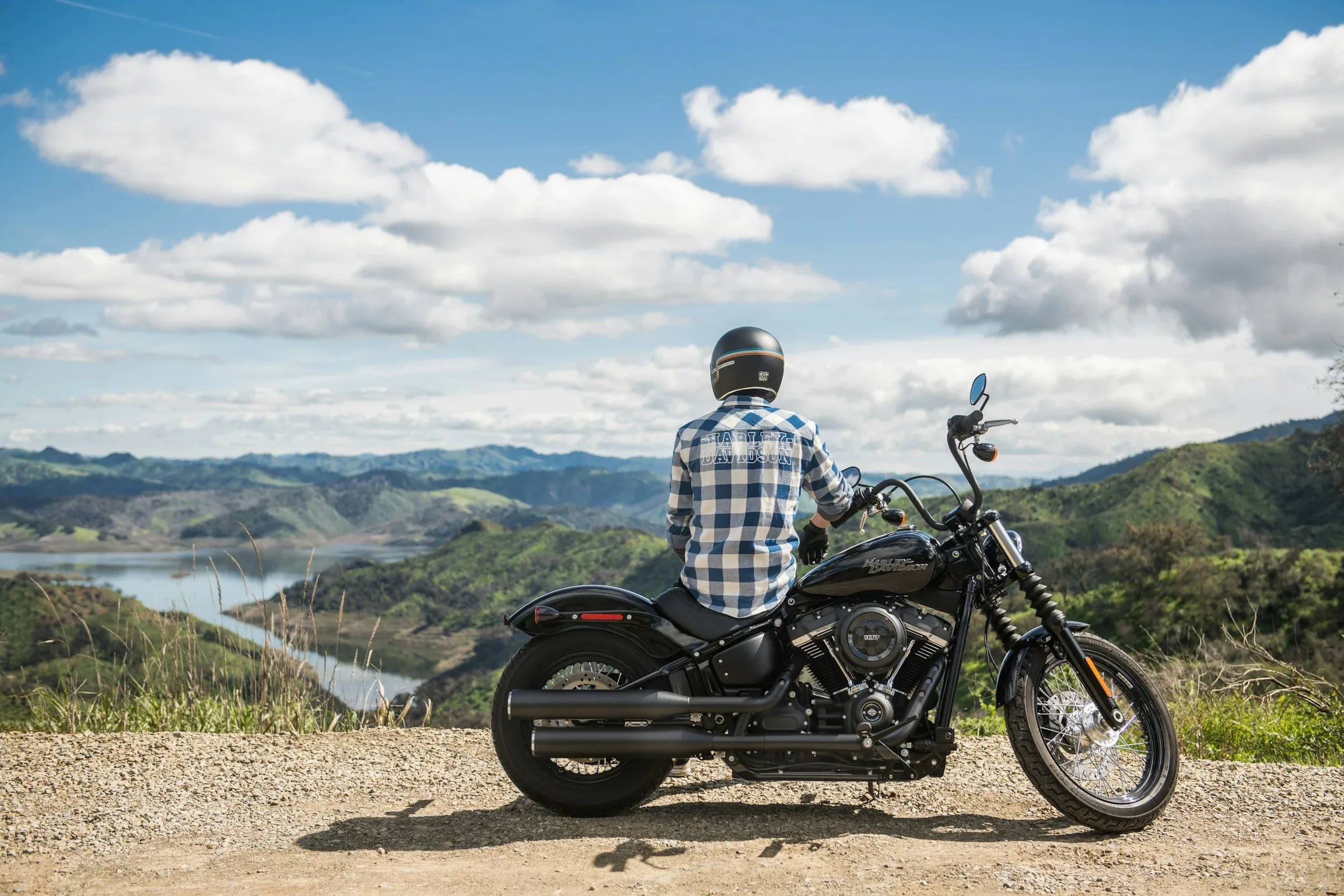 Rear view of a person wearing a helmet and plaid shirt sitting on a black Harley-Davidson motorcycle, looking out over a scenic mountain and lake landscape.