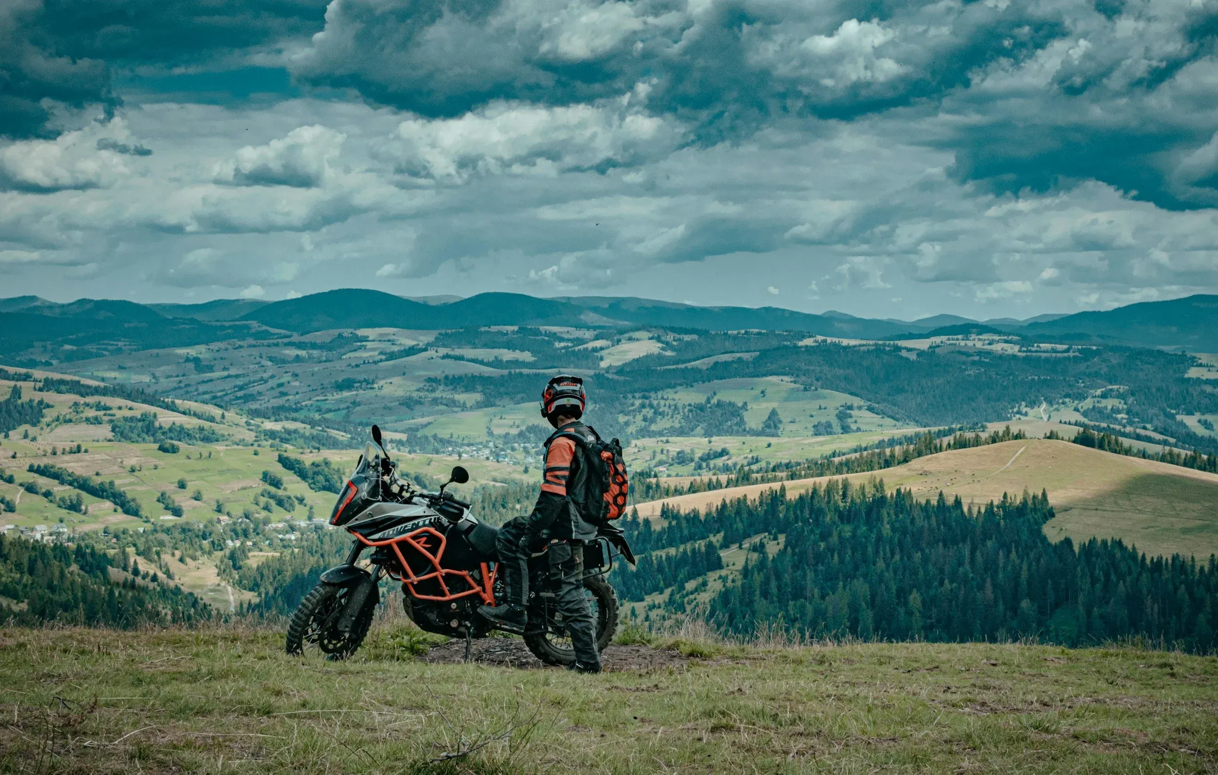An adventure motorcyclist in protective gear standing next to an orange and black KTM adventure bike on a grassy hilltop, looking out over a vast green mountain landscape under a dramatic cloudy sky.