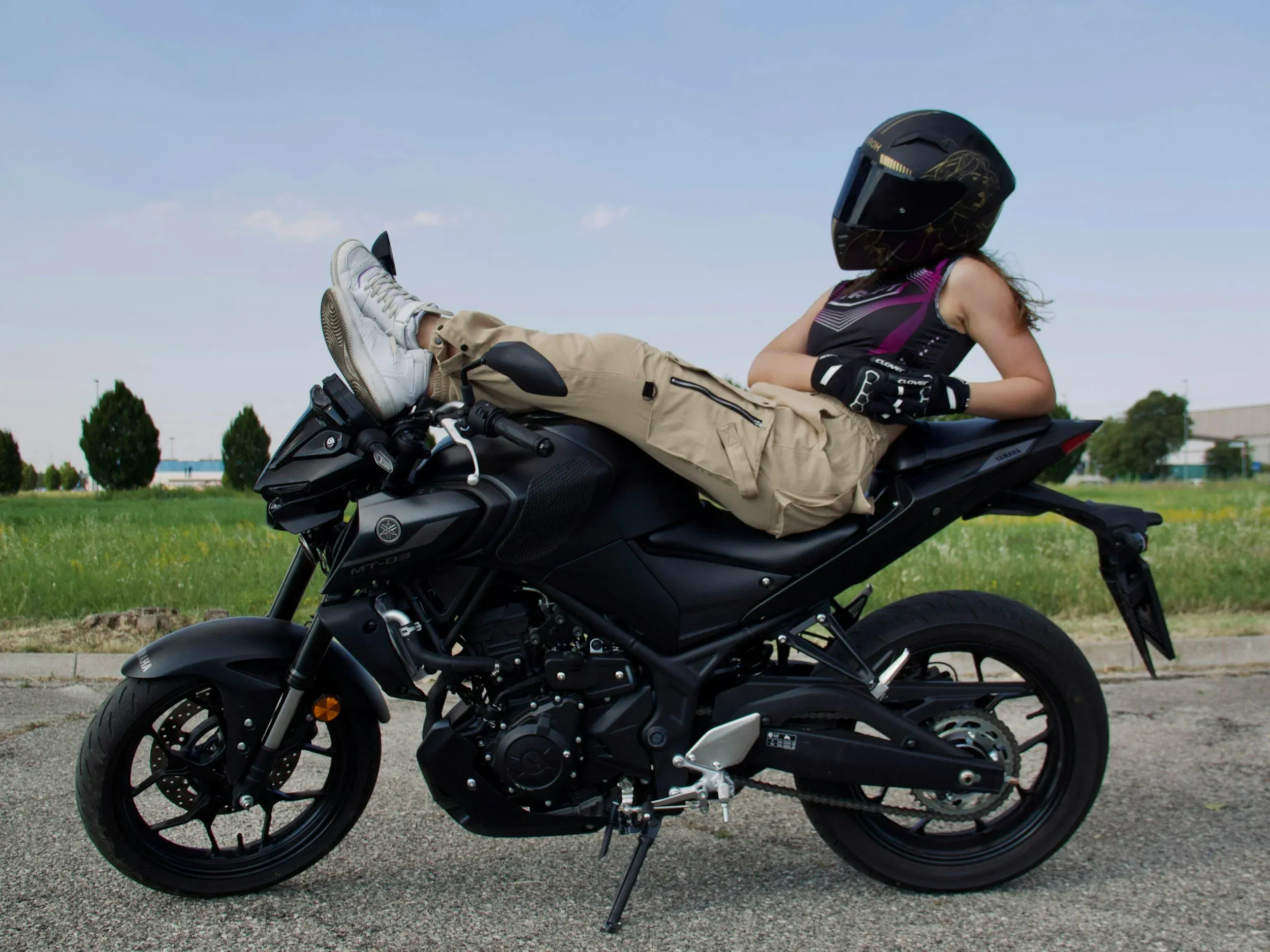 A woman wearing a helmet and cargo pants relaxes by leaning back and resting her feet on the handlebars of a black Yamaha MT-03 naked motorcycle.