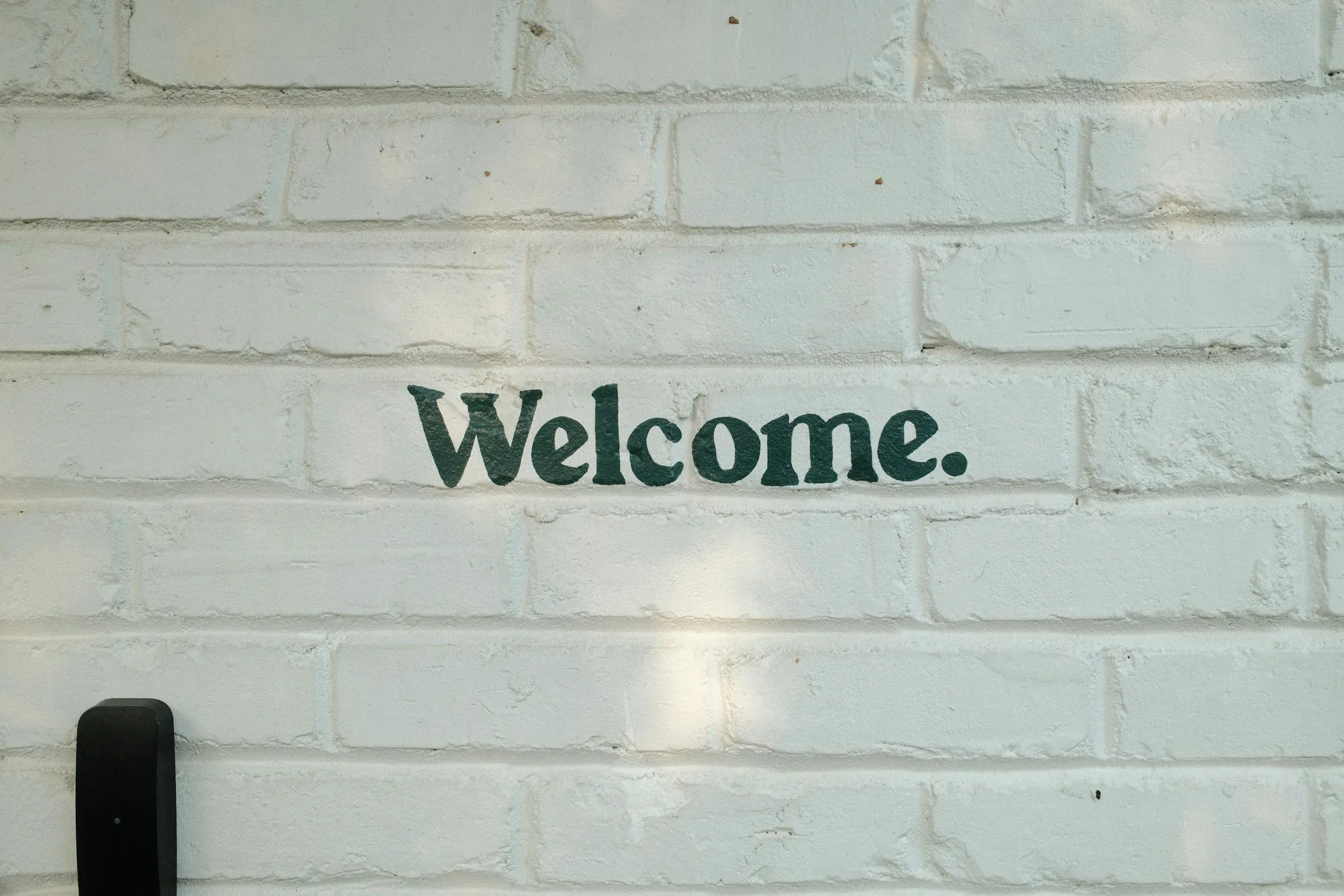 The word Welcome painted in dark green vintage block letters on a painted white brick wall with soft shadows.