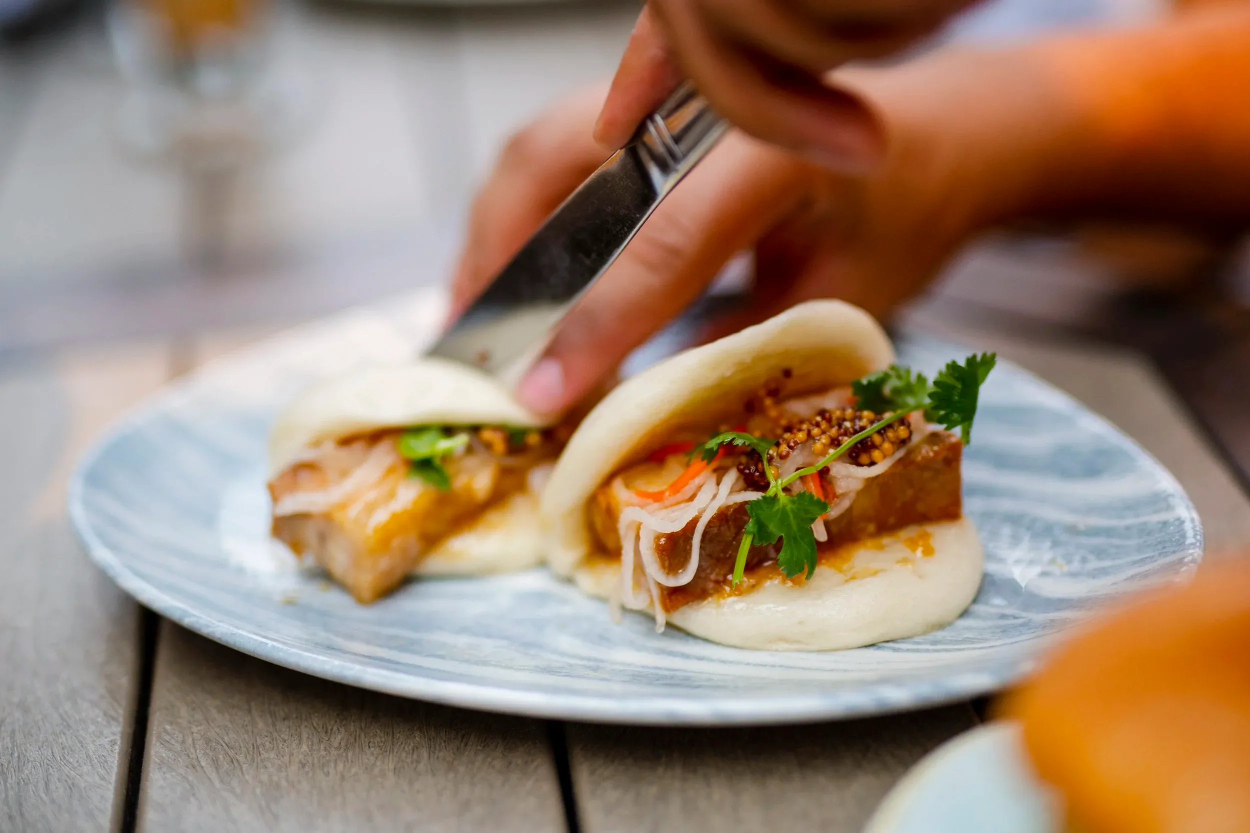 Close-up of a person using a knife to slice a gourmet pork belly bao bun garnished with mustard seeds and fresh cilantro on a blue marbled plate.