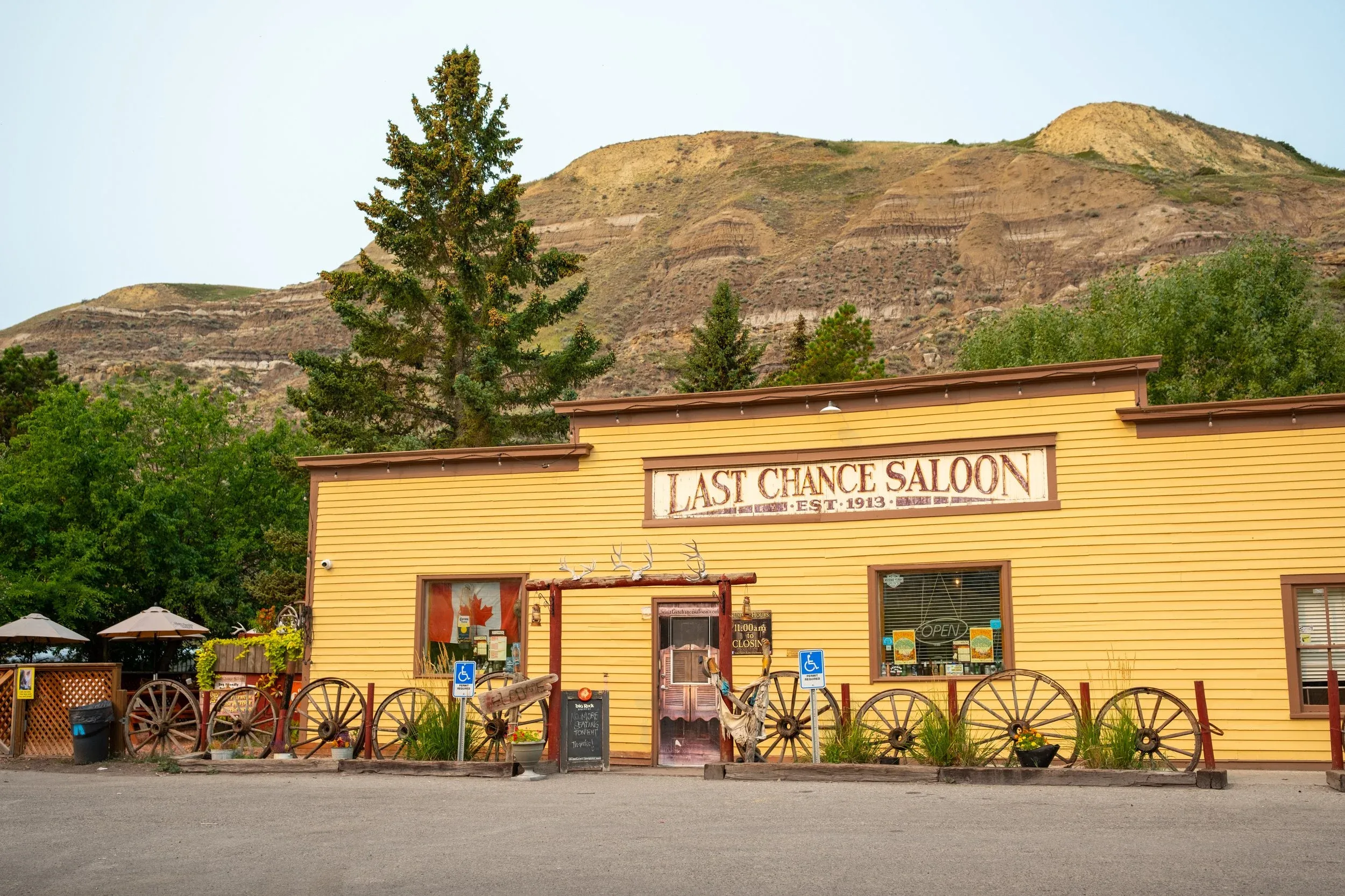 The historic yellow wooden exterior of the Last Chance Saloon with vintage wagon wheels out front, situated against desert badland hills.