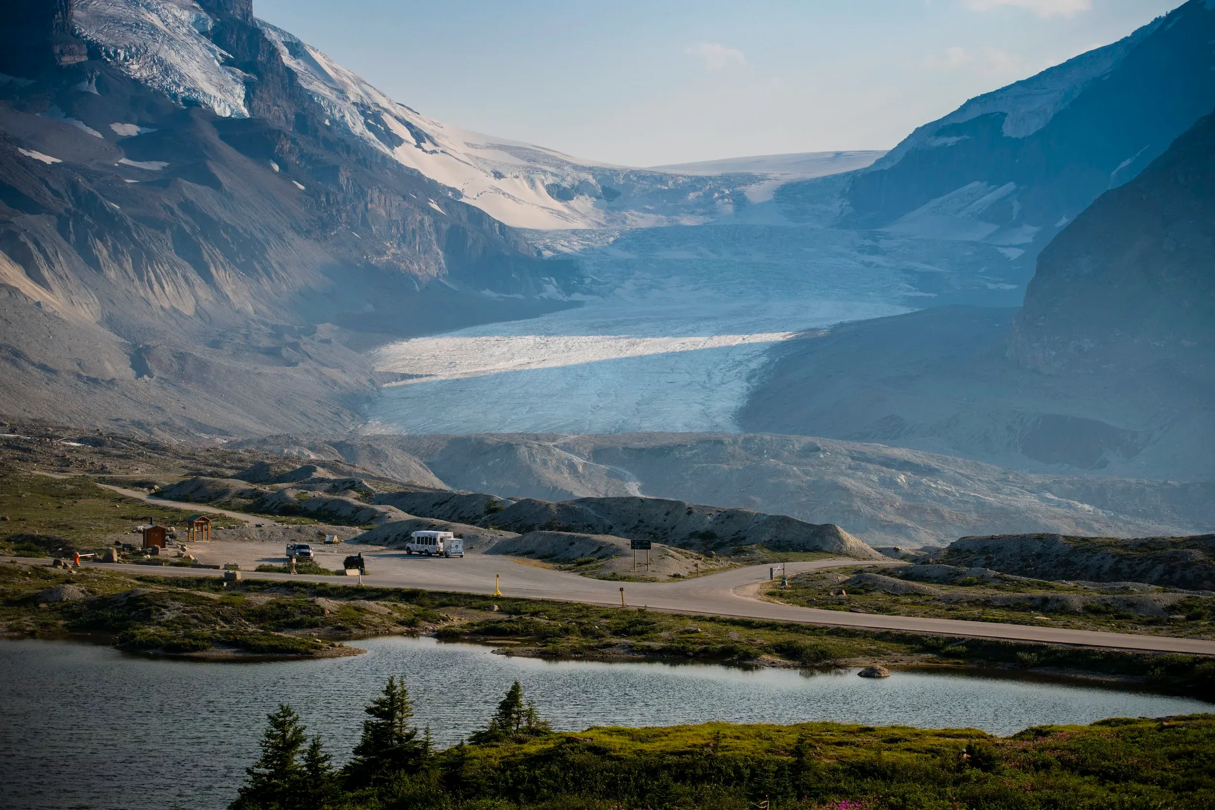 Wide landscape view of a massive alpine glacier nestled between rocky mountain peaks, with a small parking lot, road, and a calm lake in the foreground under soft daylight.