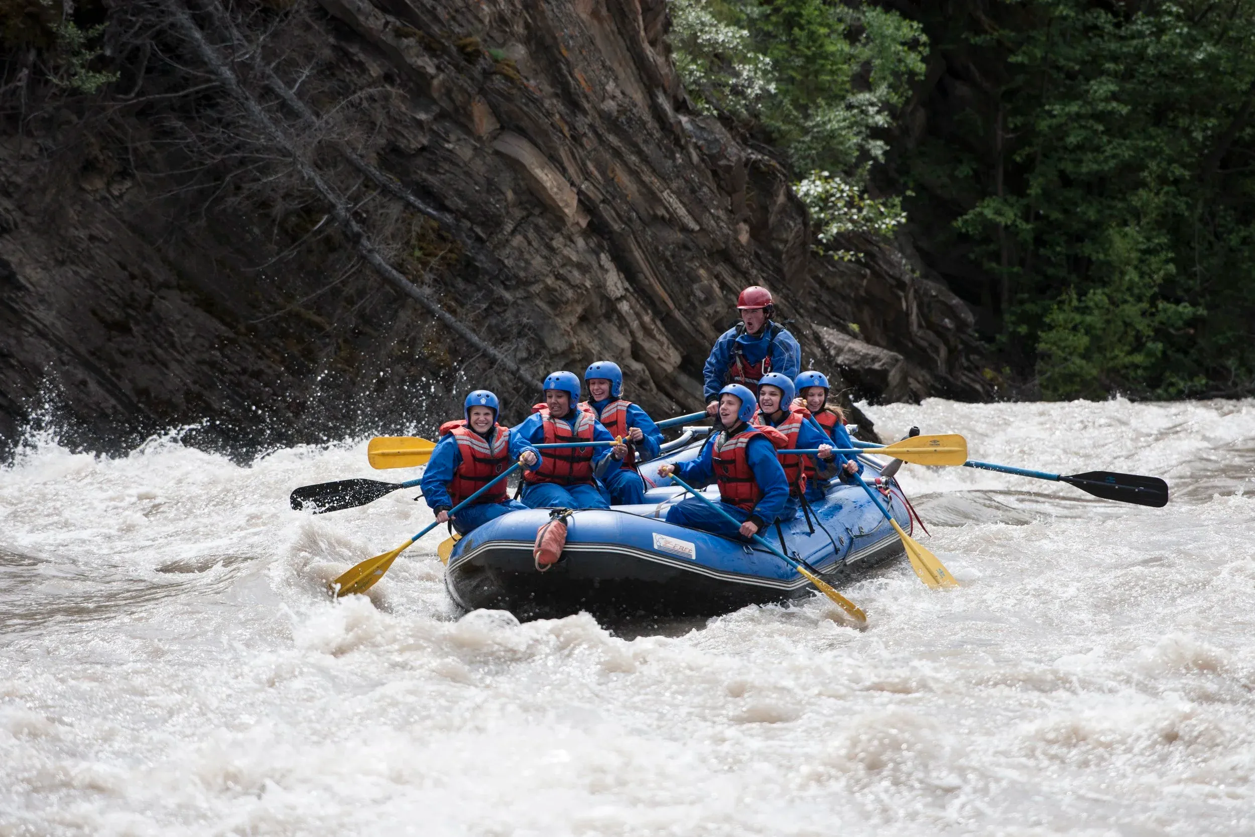 A group of people in blue helmets and red life jackets white water rafting down a rushing river alongside a steep rock wall.